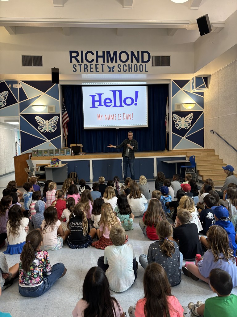 A large group of elementary students sit on the floor of a school auditorium facing the stage, where a presenter stands with a microphone. A screen behind him displays the words “Hello! My name is Dan!” under the Richmond Street School sign.