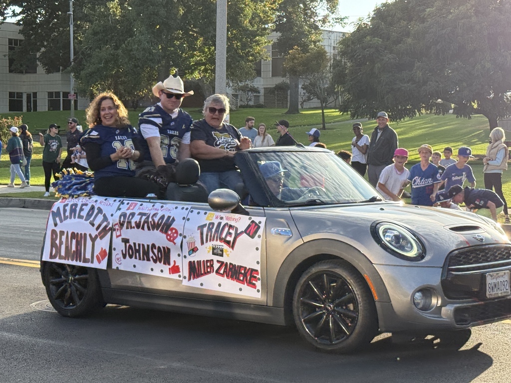 A silver Mini Cooper convertible drives in a parade with three smiling adults sitting on the back seat wearing matching blue sports jerseys. The car is decorated with a large handmade sign listing names including “Meredith Beachly,” “Dr. Jason Johnson,” “Tracey Miller Zarnikow,” and “Miller Zarnewke.” Spectators and children in team jerseys watch from the sidewalk in the background.