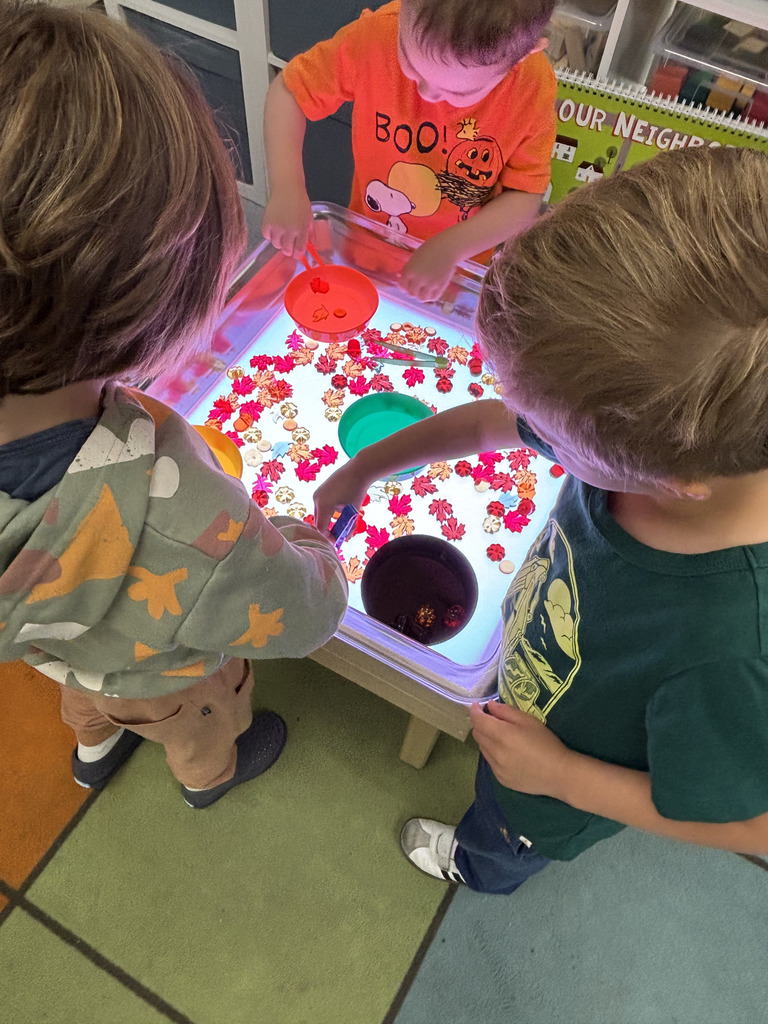 Three young children stand around a light table covered in translucent fall-themed shapes like leaves and pumpkins. They use small cups and scoops to sort the pieces into colored containers.