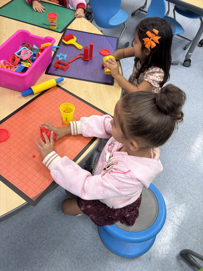 Two young children sit at a classroom table using Play-Doh and plastic tools like rollers and cookie cutters. A bright pink bin of Play-Doh tools sits in the center of the table.