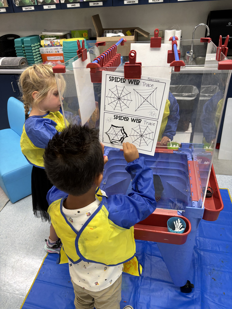 Two children wearing paint smocks stand at an easel station. One child holds a “Spider Web Trace” worksheet while the other paints or draws on clear vertical panels.