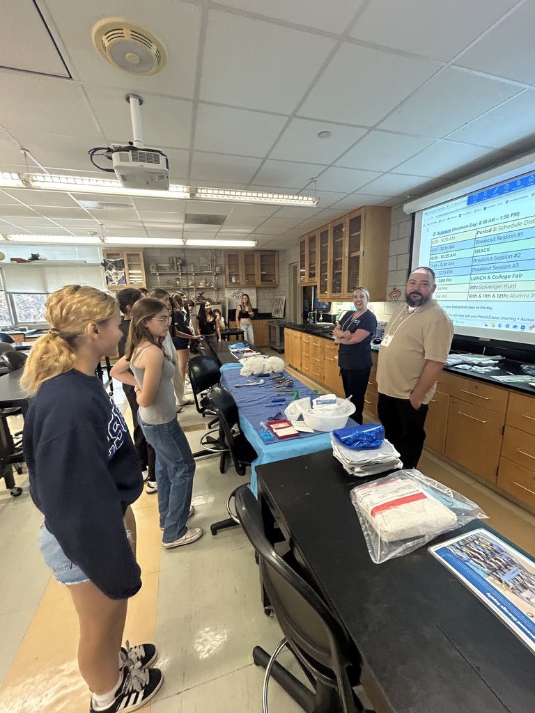 High school students stand around a lab table while two instructors at the front of the classroom lead a demonstration. The table is covered with blue cloths, medical tools, and equipment. A presentation slide with a schedule is projected onto the screen behind them.