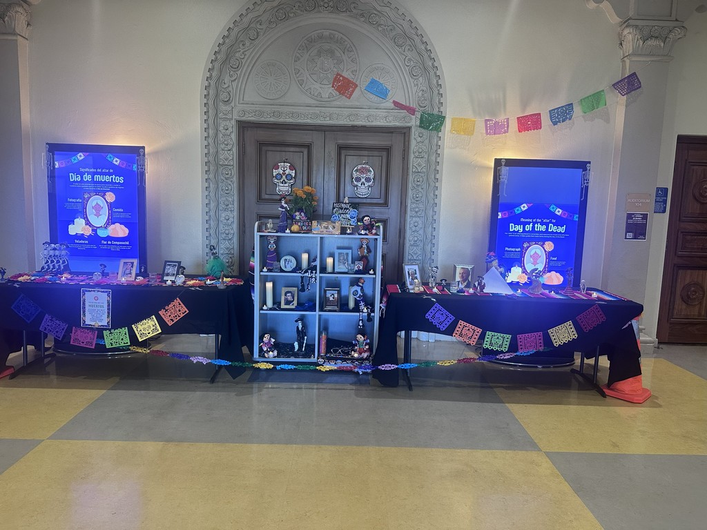 An indoor Día de Muertos (Day of the Dead) altar features tables draped with black and colorful fabric, papel picado banners, framed photos, candles, and traditional decorations. Two digital displays on either side explain the meaning of Día de Muertos in Spanish and English.