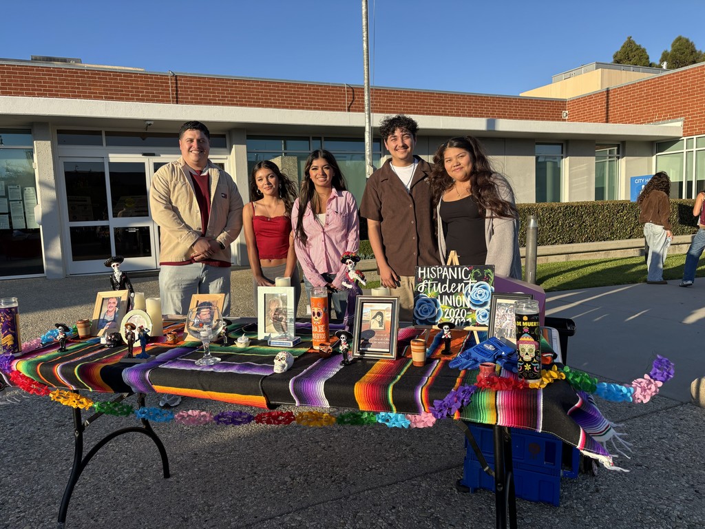 Five people stand smiling behind a colorful Day of the Dead display table outdoors. The table is covered with a striped blanket and decorated with framed photos, candles, small skeleton figurines, and flowers. A small sign reads “Hispanic Student Union.”