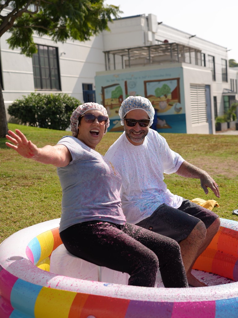 Two staff members sit in a colorful inflatable pool, laughing and waving at the camera while covered in splattered foam. Both wear shower caps and sunglasses during a sunny outdoor school event.