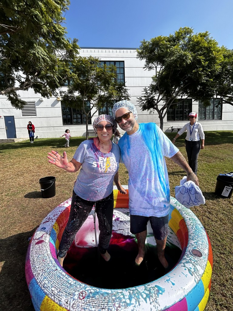Two smiling staff members, covered in colorful foam, stand inside a rainbow inflatable pool after a playful soaking. Both wear shower caps and casual clothes, enjoying the outdoor event under sunny skies.