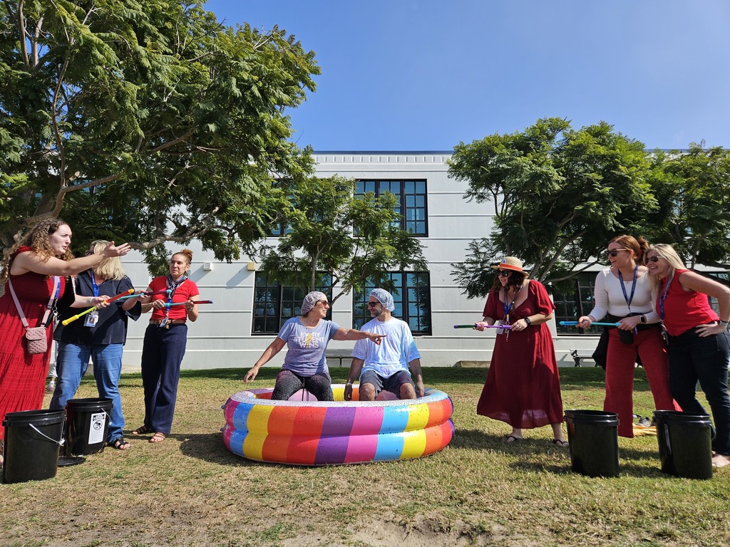 A group of teachers stand outdoors on a sunny day, laughing and cheering as two colleagues sit in a colorful inflatable pool. The seated participants wear shower caps and are about to be sprayed with water or foam as part of a fun school activity.