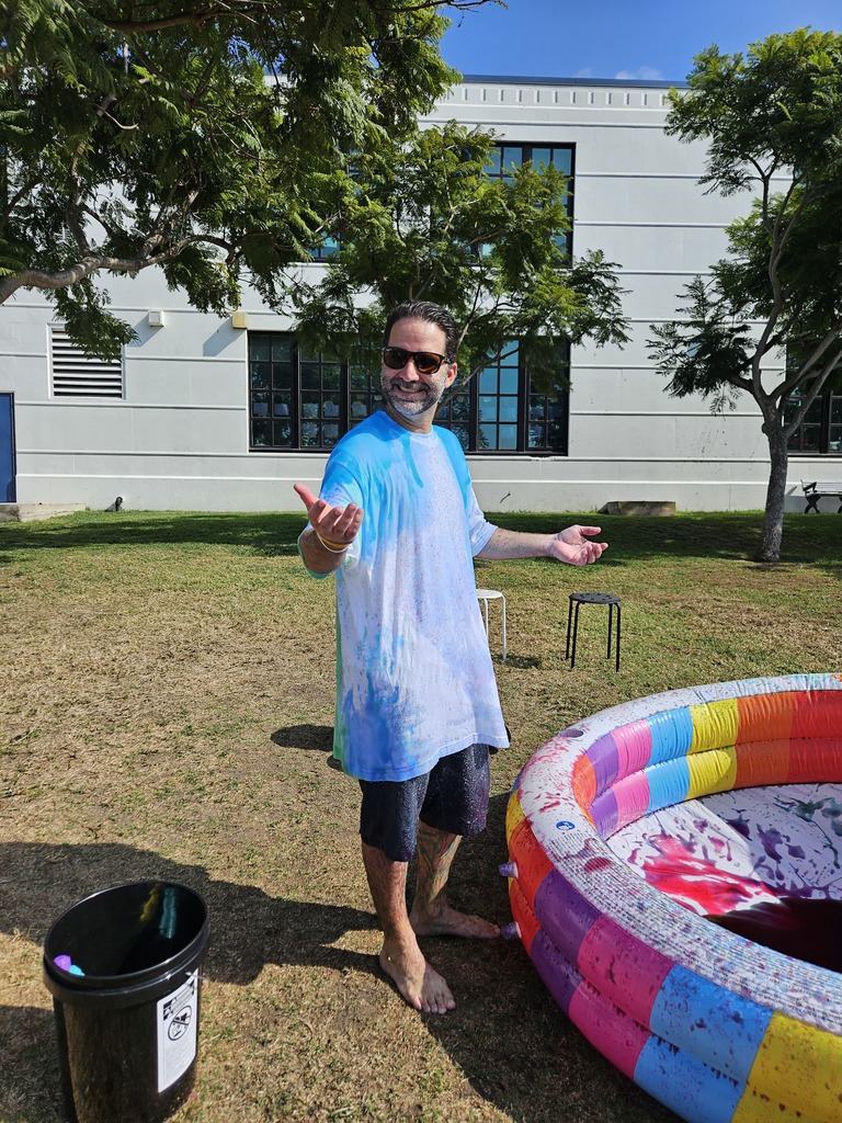 A staff member, smiling and covered in blue and pink foam, stands barefoot beside a rainbow inflatable pool during an outdoor school event. Buckets and trees are visible in the background.