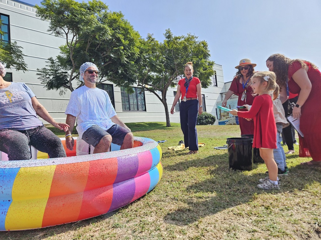 A child and several adults laugh as they prepare to spray two staff members sitting in a rainbow inflatable pool. The participants wear protective caps and shirts while others watch and smile in the background.