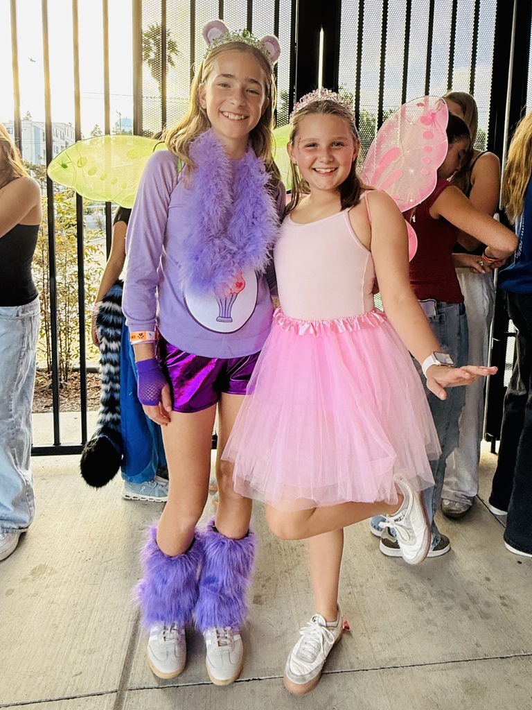 Two students in fairy costumes smile for a picture—one dressed in purple with fuzzy boots and wings, and the other in a pink tutu and wings.