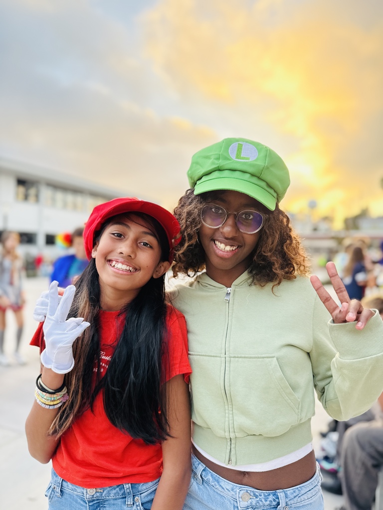 Two students dressed as Mario and Luigi pose outdoors at sunset, each flashing a peace sign and smiling at the camera.