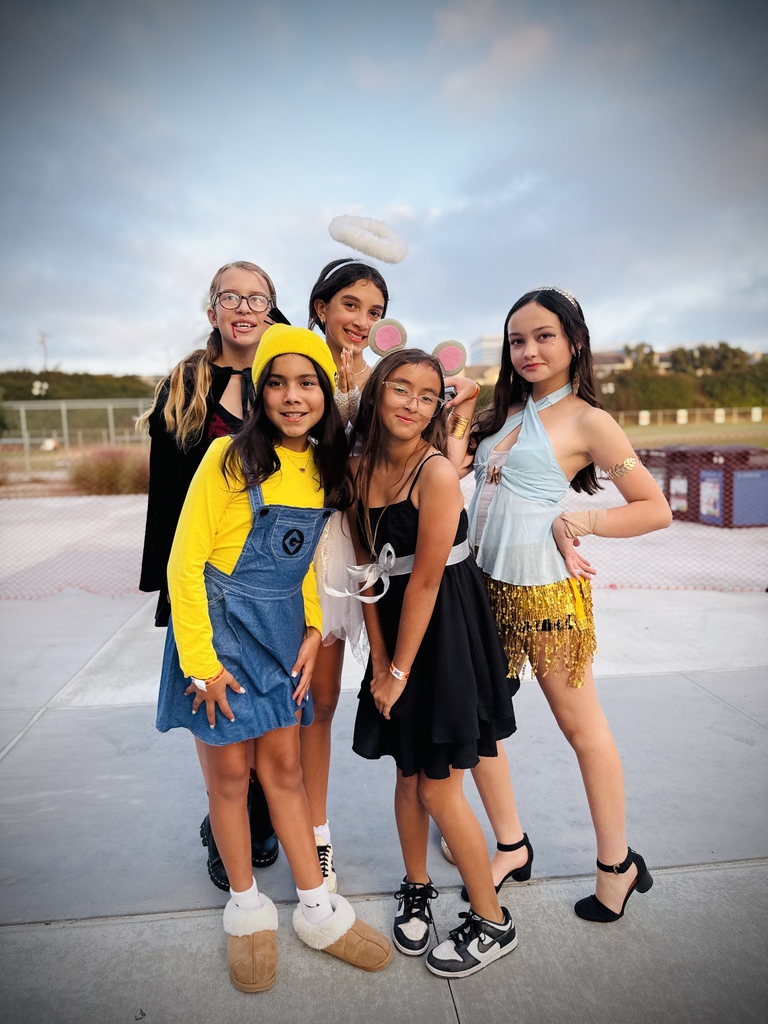 Five students pose outdoors in various costumes, including a minion, an angel, a mouse, and a 1920s-inspired outfit, smiling under a cloudy evening sky.