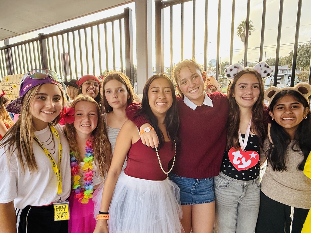 A group of students in colorful costumes smile together at a Halloween-themed event. Their outfits include animal ears, a tutu, a lei, and creative accessories.