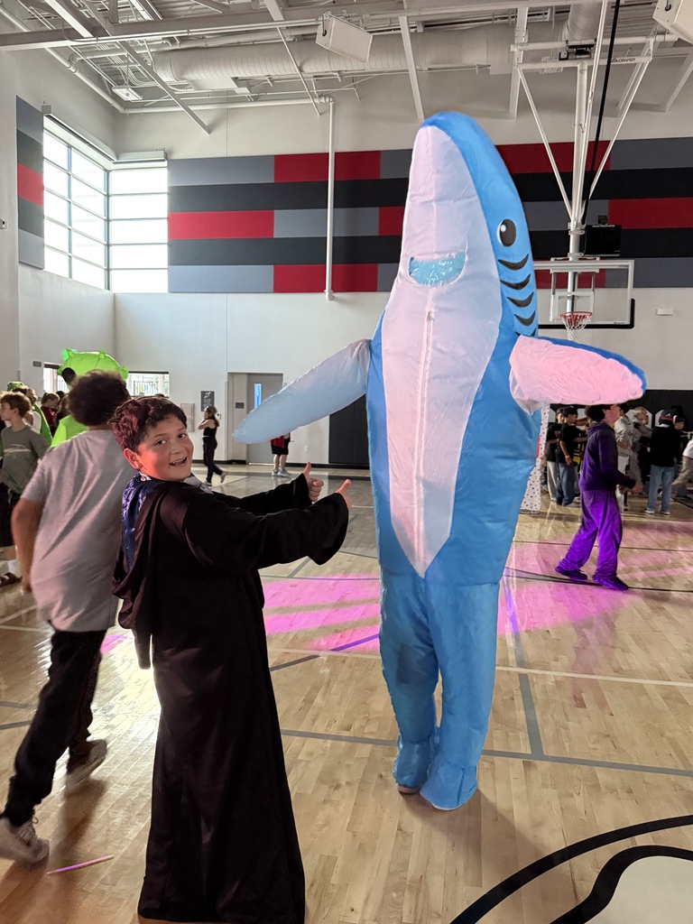 A student dressed in a dark robe gives a thumbs-up to someone wearing a large inflatable shark costume inside a school gymnasium during a Halloween event.
