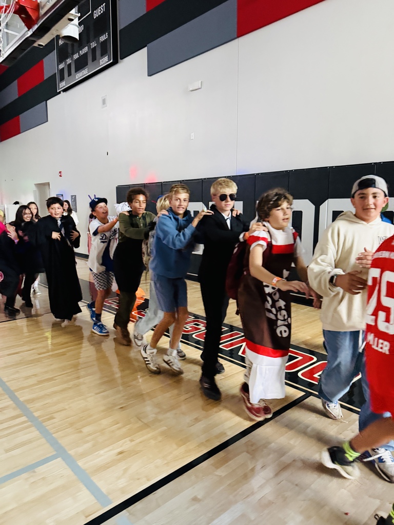 A line of students in costumes, including a person in sunglasses and a candy bar costume, form a conga line inside a gym decorated in red, gray, and black.