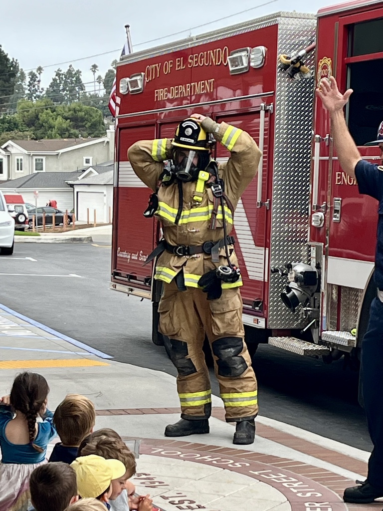 A firefighter wearing full turnout gear, helmet, and breathing apparatus demonstrates fire safety equipment to a group of seated students.