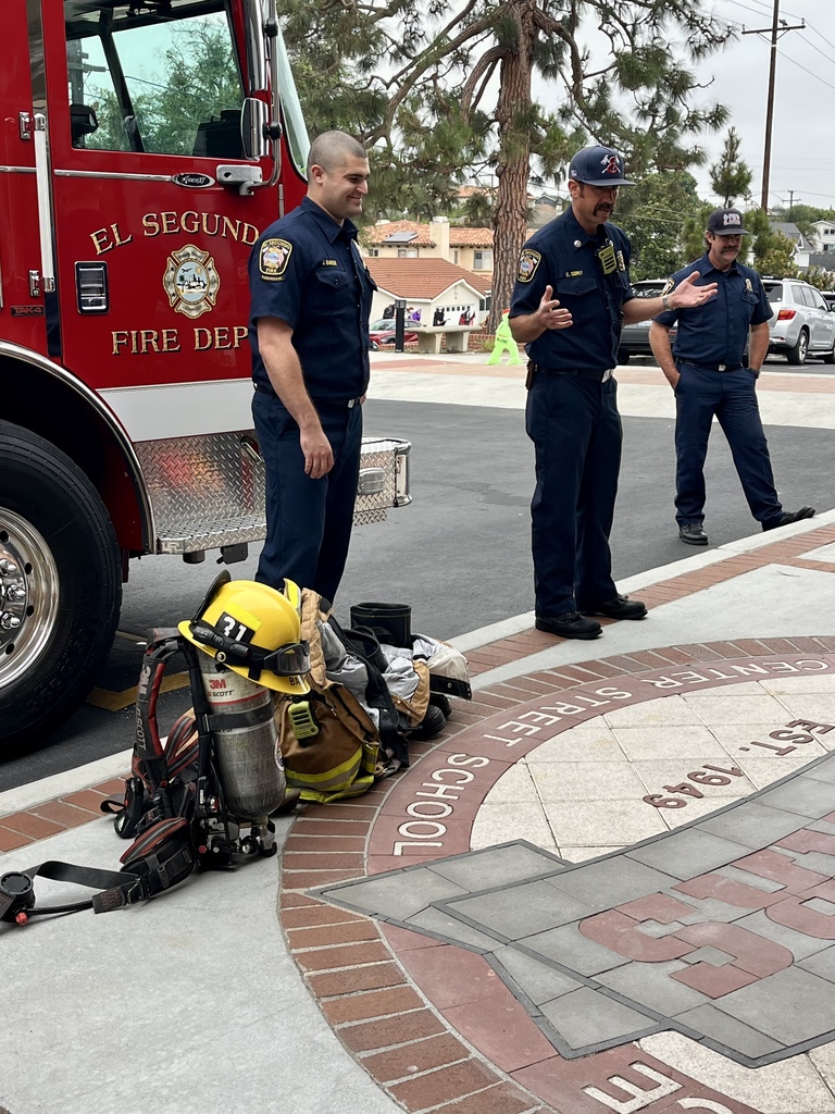 Three firefighters stand in front of a red El Segundo Fire Department truck, speaking to students outside.
