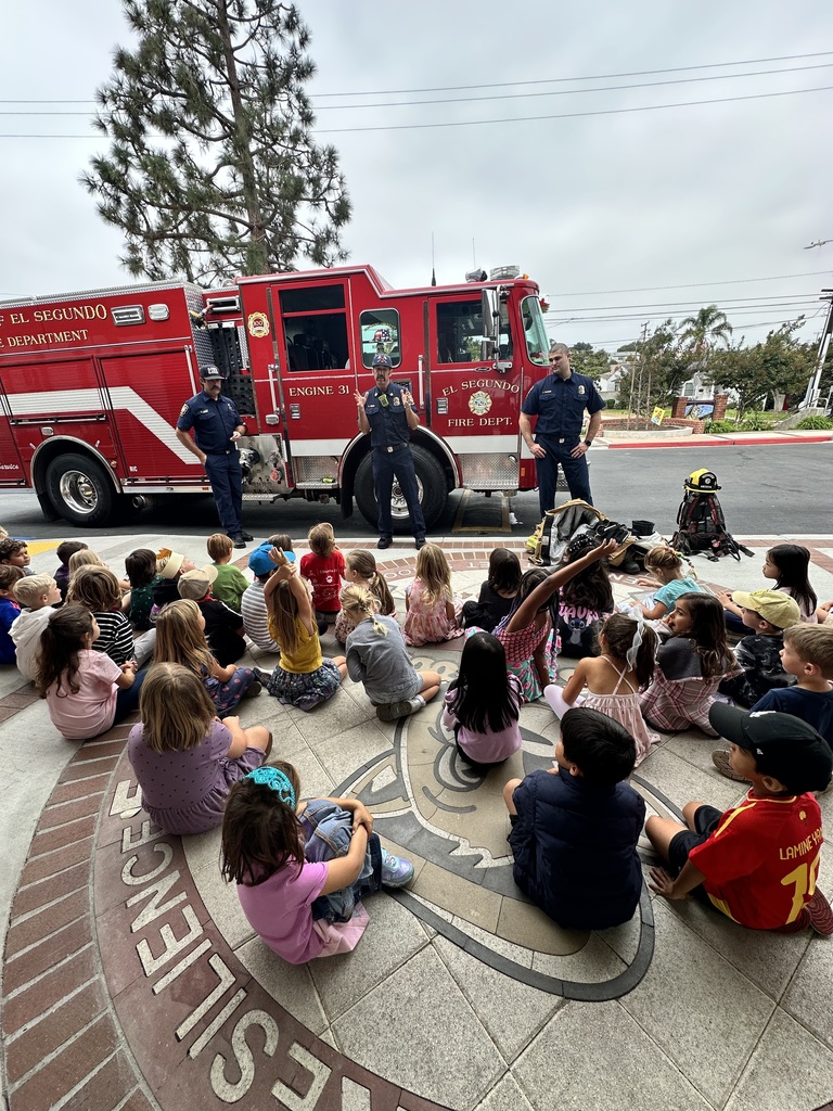 A group of young students sit cross-legged on a circular school courtyard emblem while three firefighters stand in front of a bright red El Segundo Fire Department truck.
