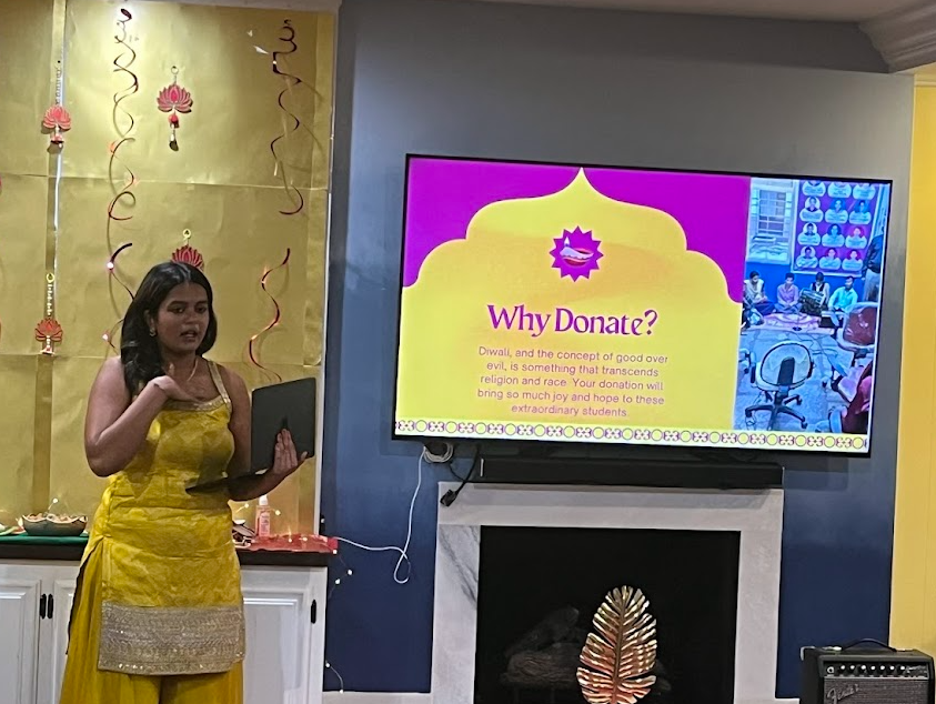 A woman wearing a bright yellow traditional outfit presents in front of a decorated wall and a large screen. The screen displays a slide with the heading “Why Donate?” and text about Diwali, good over evil, and bringing joy to students. The setting appears festive with gold and red decorations.