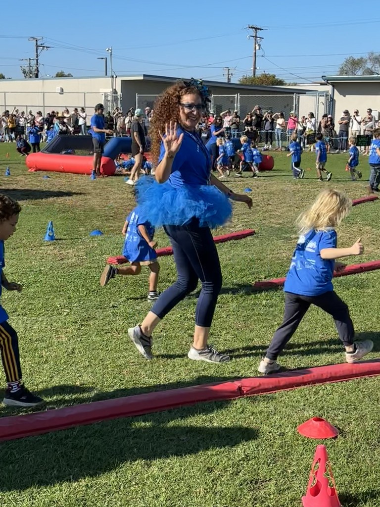 A woman in a blue shirt, blue tutu, and fun accessories waves while walking alongside young children participating in the Fall Flutter