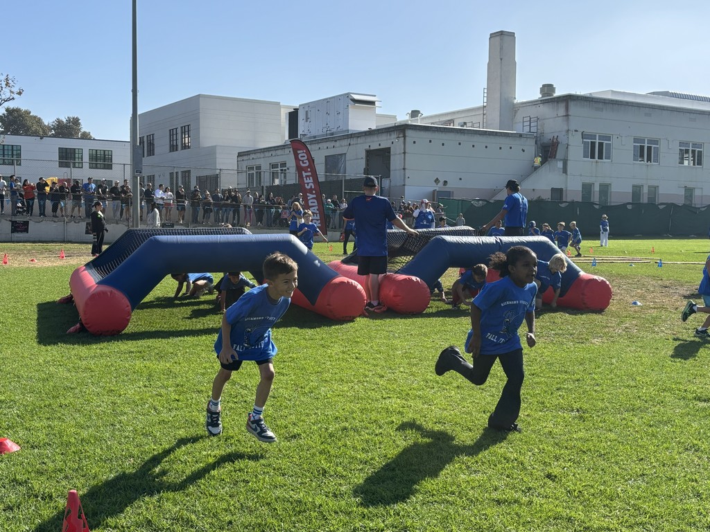Young children in blue shirts run past an inflatable obstacle while other students crawl underneath it