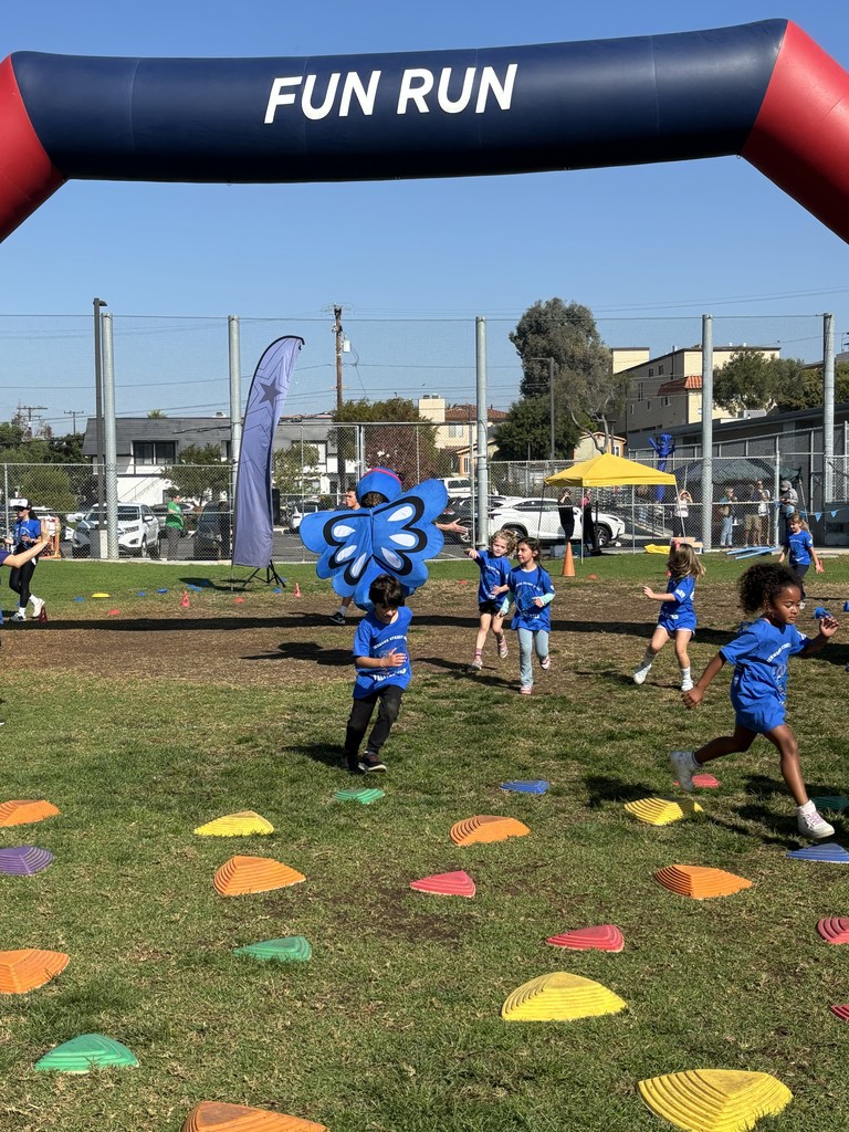 Children in blue shirts run across a grassy field filled with colorful stepping-stone markers under a large inflatable arch labeled “FUN RUN.”