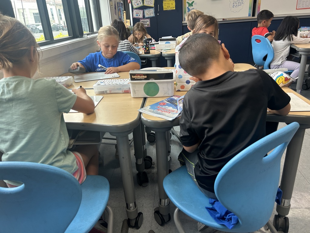 Elementary students sit at grouped desks working on individual assignments. Some write on papers or whiteboards while others focus on classroom materials.