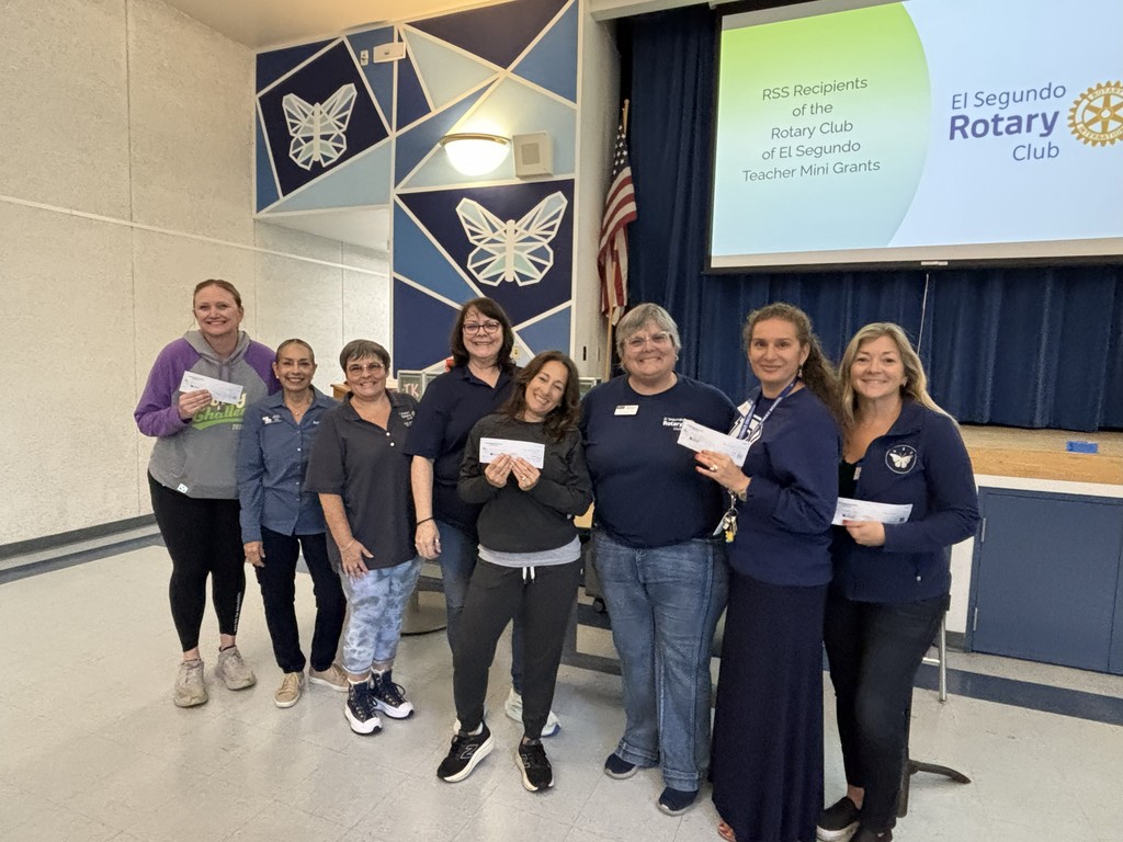 A group of eight staff members stand together in a school multipurpose room, smiling and holding Rotary Club mini-grant checks. A projector screen behind them displays the text “RSS Recipients of the Rotary Club of El Segundo Teacher Mini Grants” alongside the Rotary Club logo.