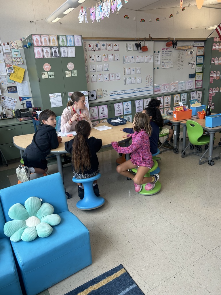 A teacher sits at a small group table with five young students gathered around her in a colorful elementary classroom.