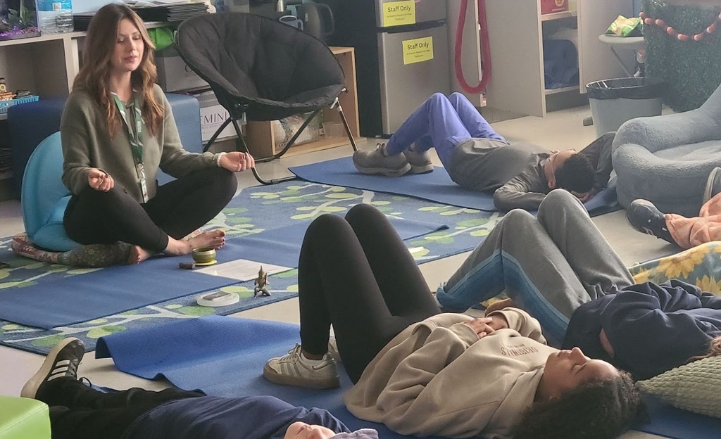A teacher sits cross-legged on a cushion at the front of the room leading a mindfulness or relaxation activity. Several students lie on yoga mats on the floor with eyes closed, participating in the exercise.