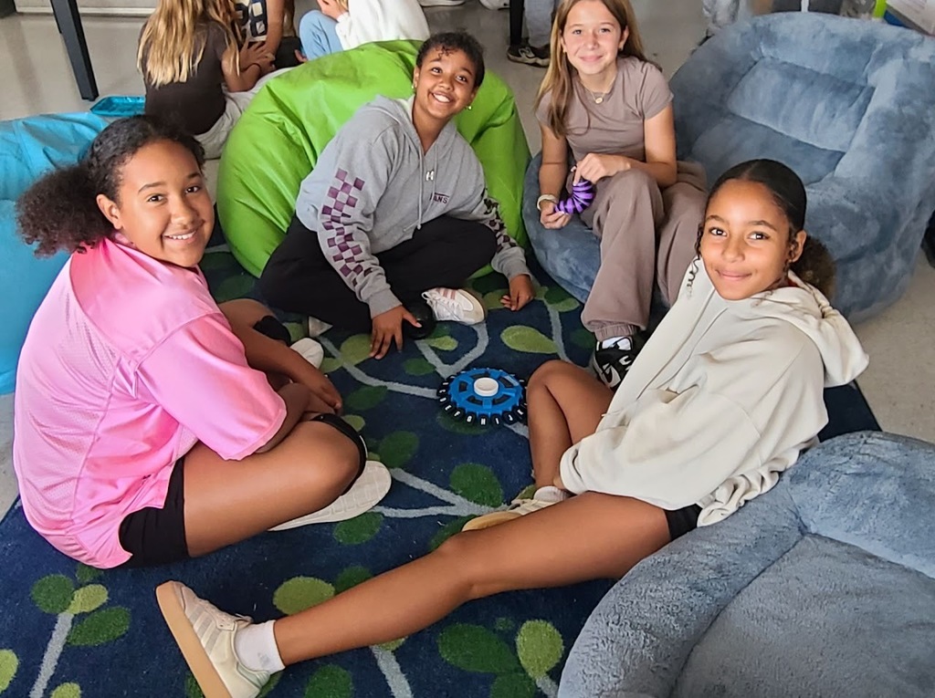 Four girls sit together on a patterned blue and green rug in a classroom. They are smiling at the camera while sitting around a small blue game spinner.