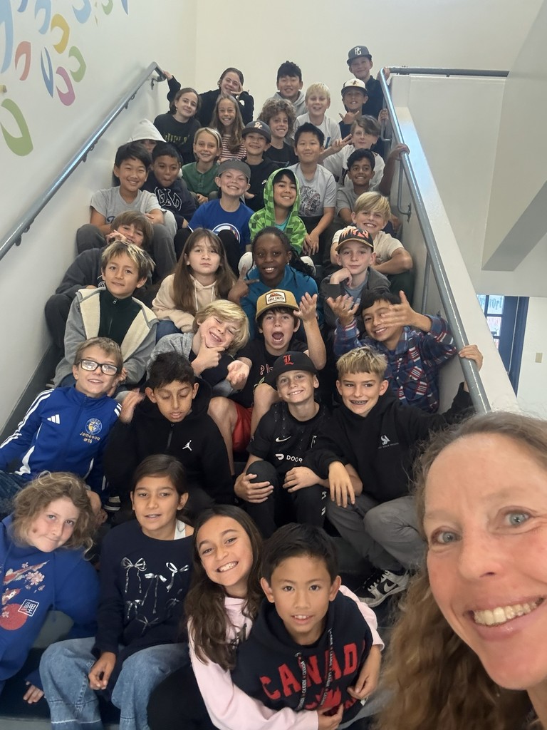 A large group of elementary students sits together on a staircase, smiling and posing for a photo. They fill the steps from top to bottom, with a teacher taking a selfie from the lower right corner.