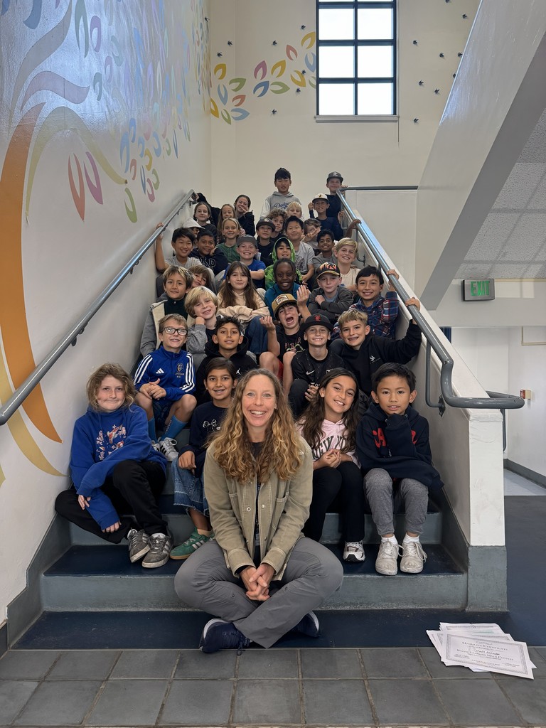 A teacher sits cross-legged at the bottom of a staircase, smiling, with a large group of elementary students seated on the steps behind her