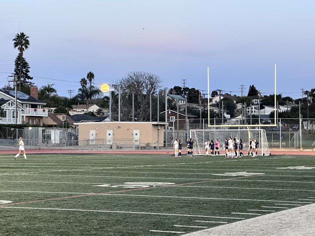 A girls’ soccer game takes place on a turf field at dusk. Most of the players gather near the goal on the right side of the image, while one player walks alone on the left.