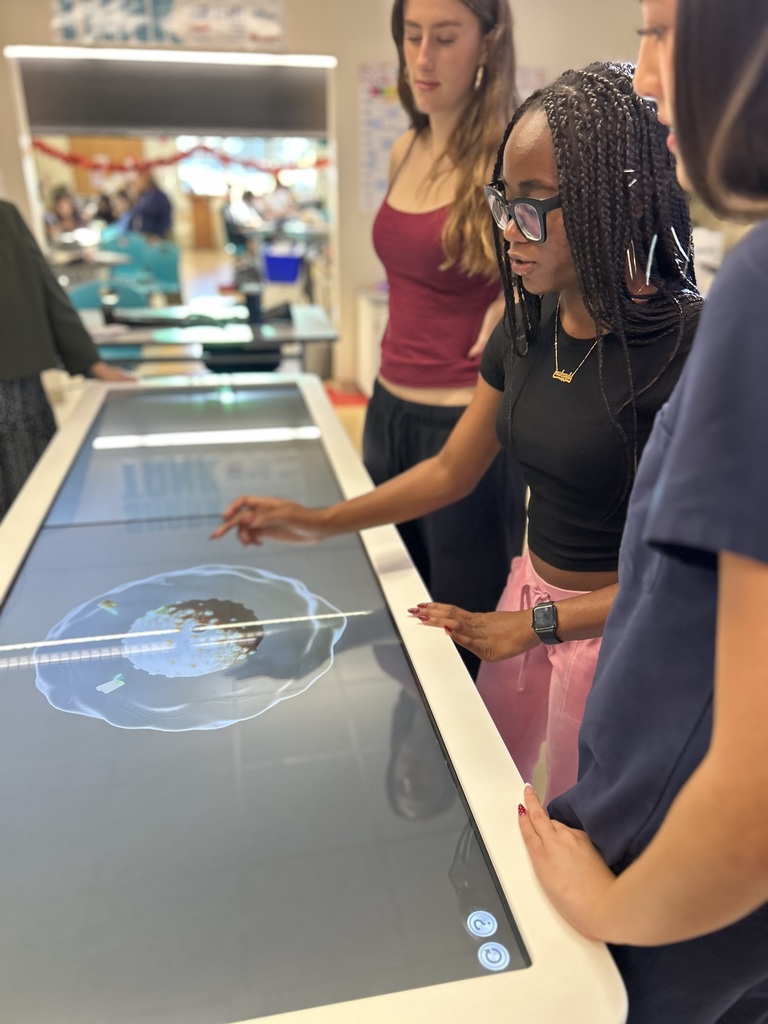 Students interact with a large touchscreen table displaying a 3D anatomical model in a classroom or lab setting.