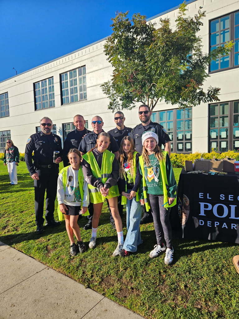 A group of El Segundo police officers stands with smiling students in safety vests in front of a school during an outdoor event.