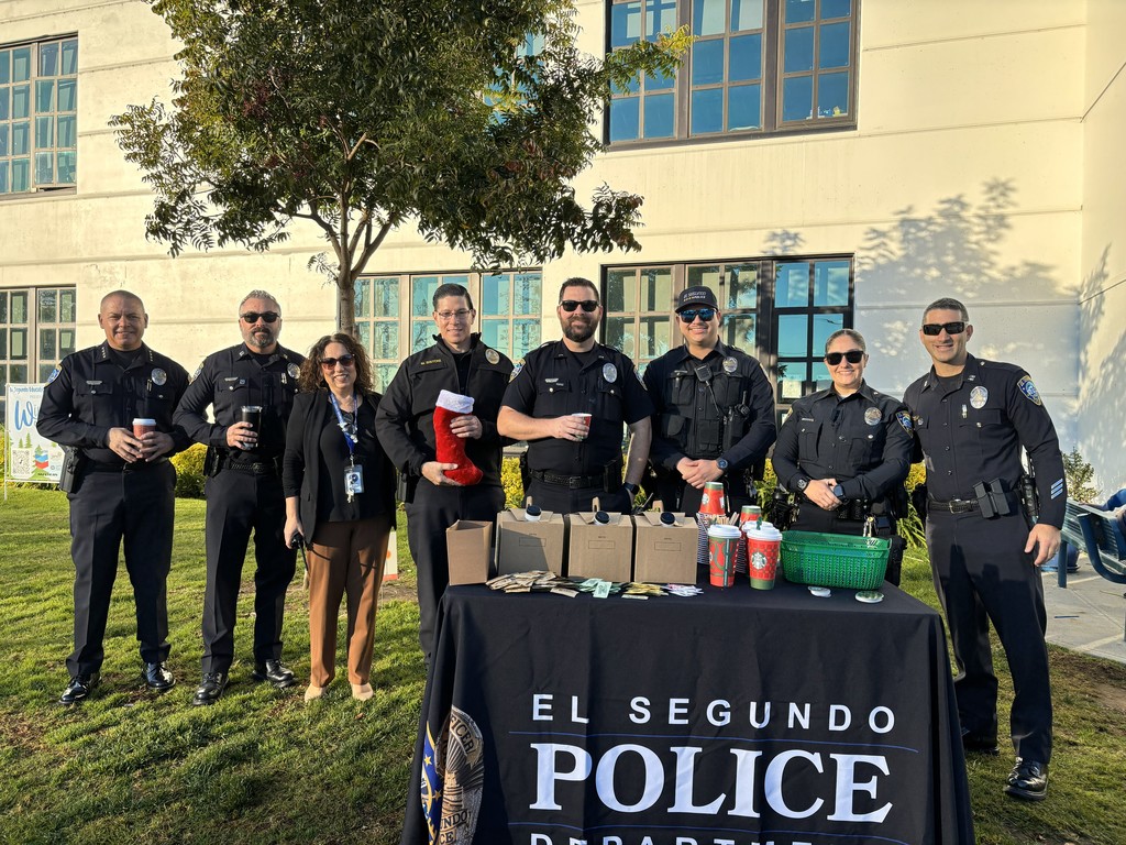El Segundo police officers and a school staff member pose smiling behind a table with coffee and giveaway items outside a school.