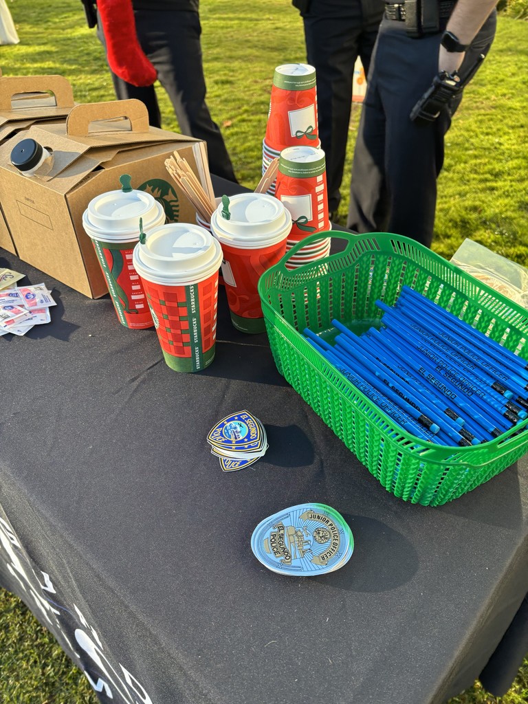 Close-up of a table with Starbucks coffee, police-themed pencils, stickers, and “Junior Detective” badges at a community event.