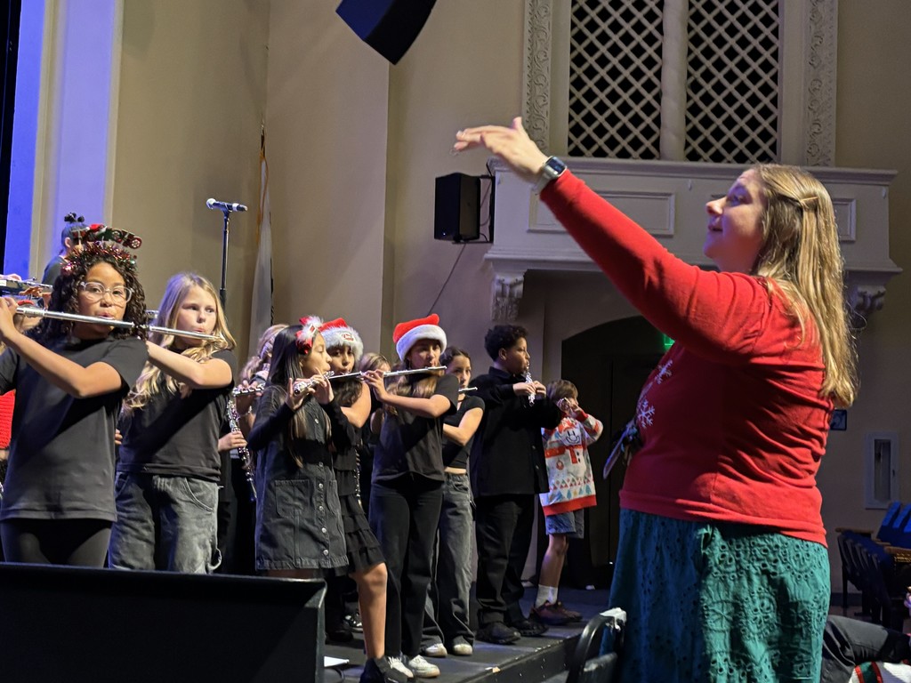 A teacher in holiday attire conducts a group of flute players, many wearing Santa hats or reindeer antlers, during a concert.