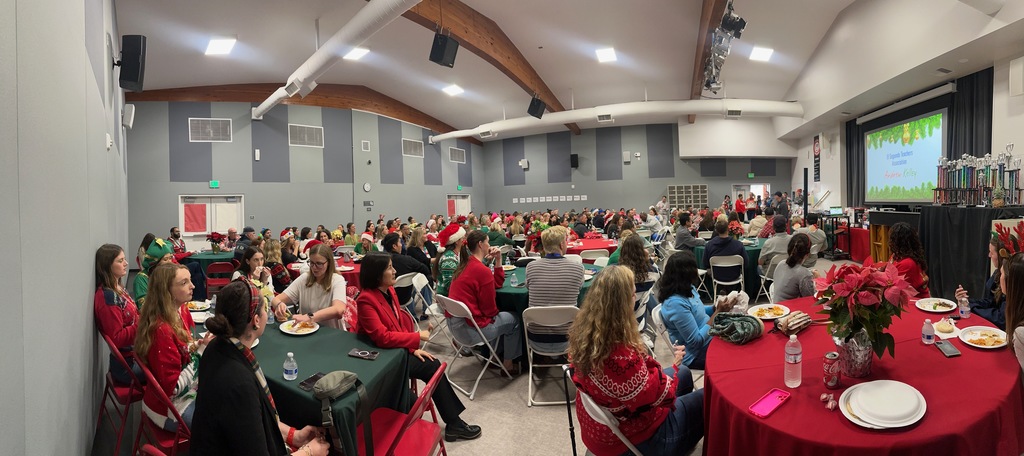 A large group of staff and guests, many in festive holiday attire, are seated at round tables enjoying a meal and presentation in a decorated auditorium.
