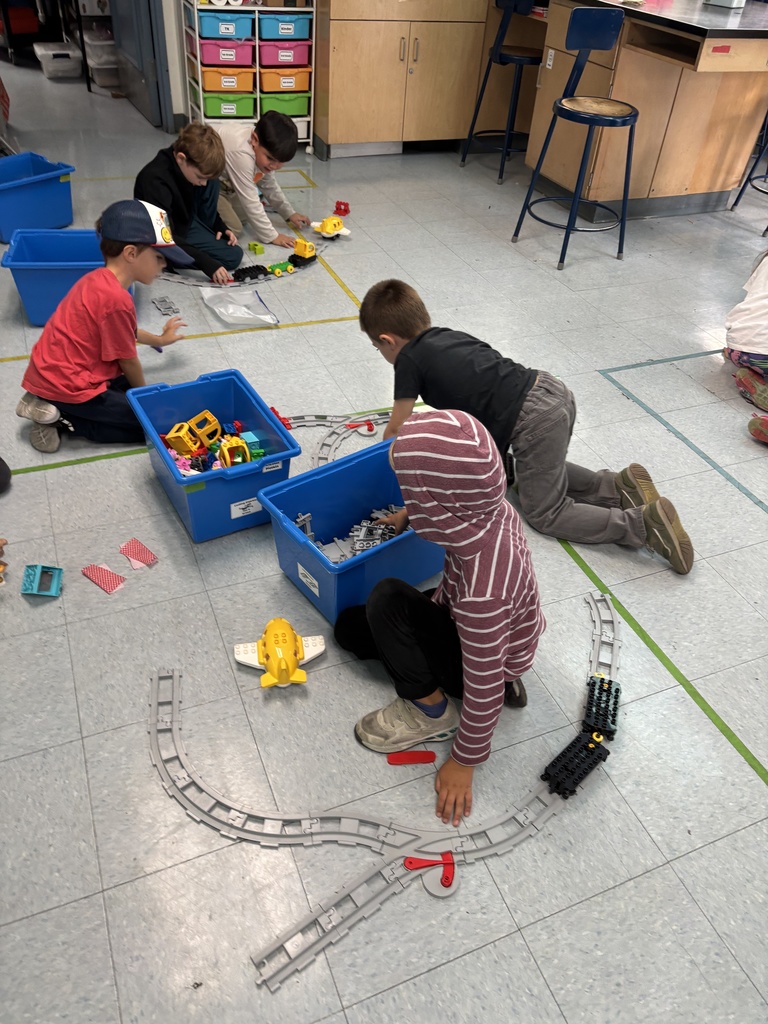 Young students build and play with plastic train tracks and toy vehicles on the classroom floor using blue bins of parts.