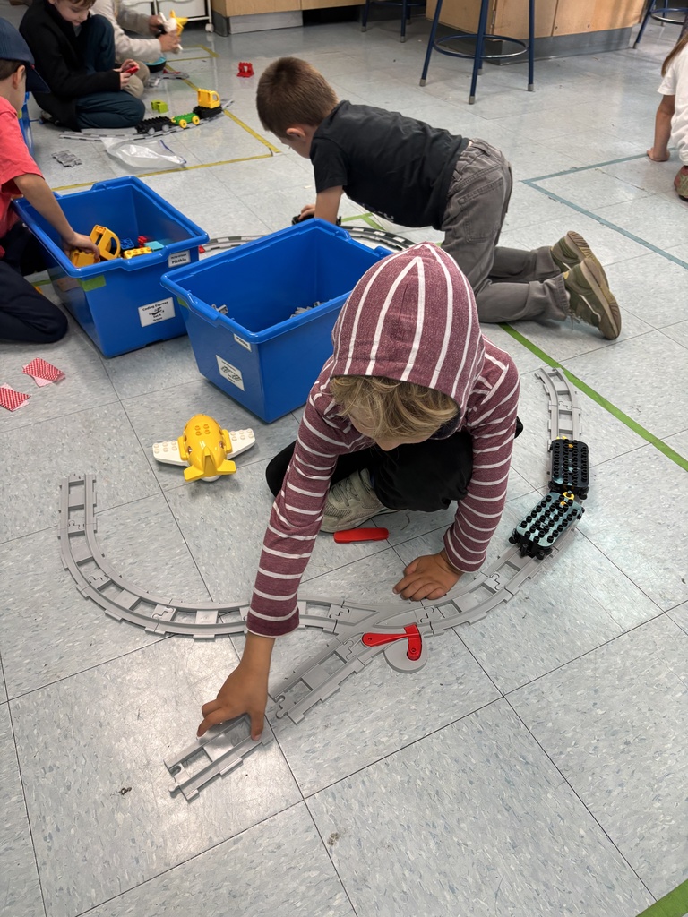 A child in a striped hoodie connects plastic train tracks on the floor while classmates build nearby with construction toys.