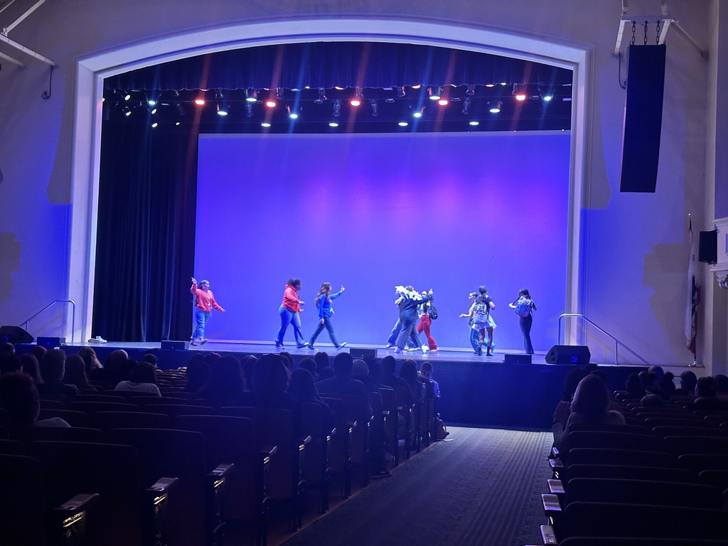 Dancers continue their routine on stage, mid-movement, as audience members watch in a dimly lit auditorium.