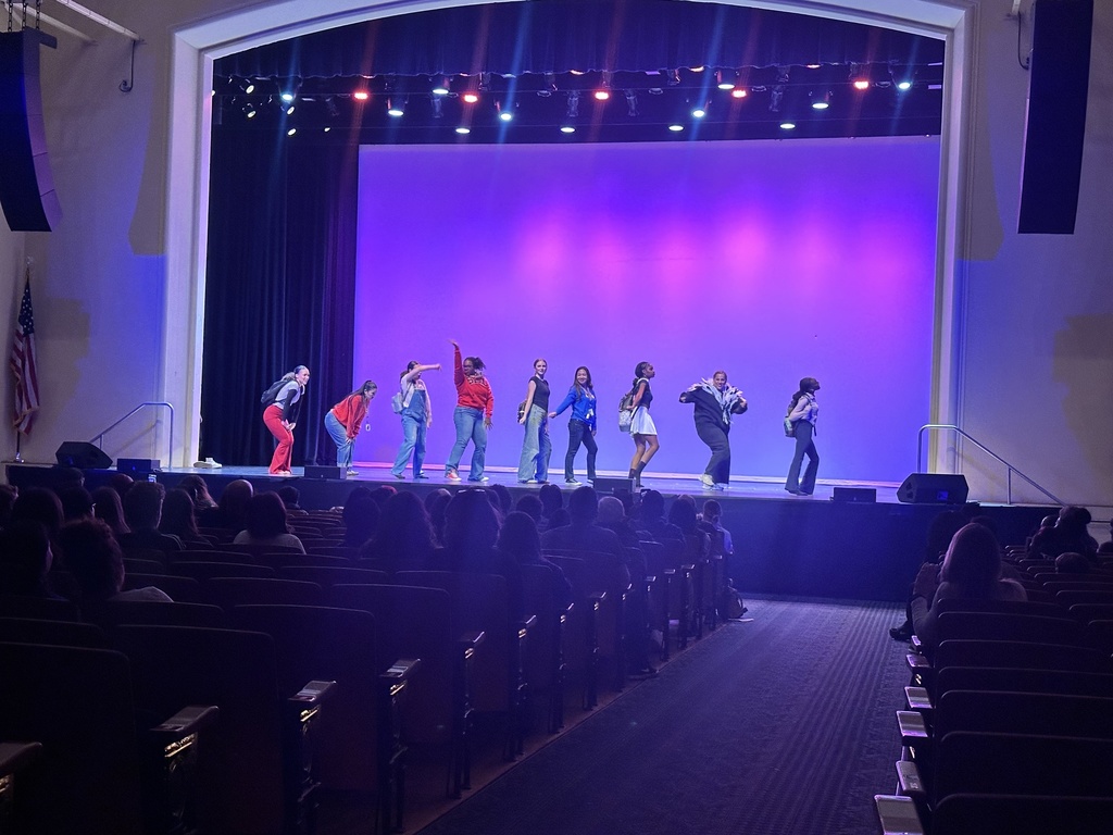 A group of performers in colorful outfits dances on stage under purple and blue lighting