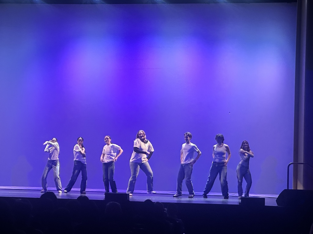 Eight dancers in white tops and jeans pose mid-performance on stage, lit with a cool purple and blue backdrop.