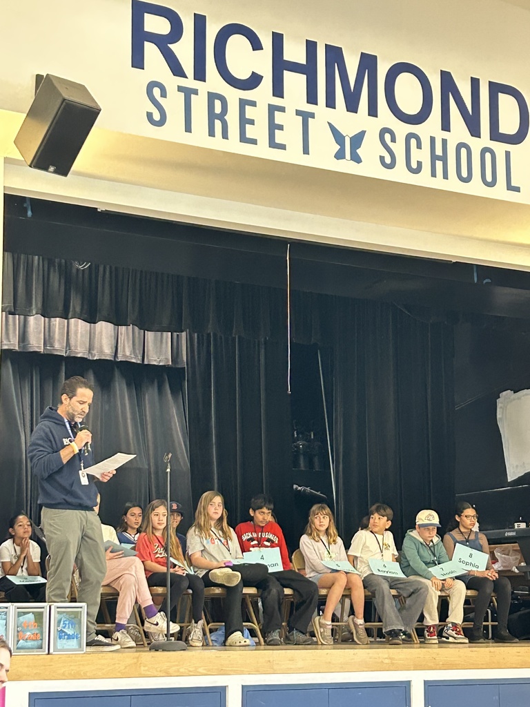 A spelling bee is in progress on the Richmond Street School stage, with a teacher reading words and student participants seated with number tags.