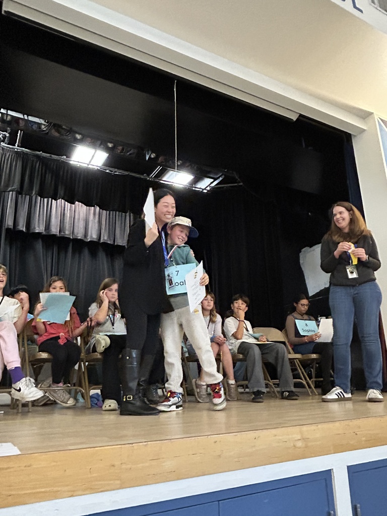 A student wearing a medal and holding a certificate smiles while being congratulated by a teacher on stage, with peers seated in the background.