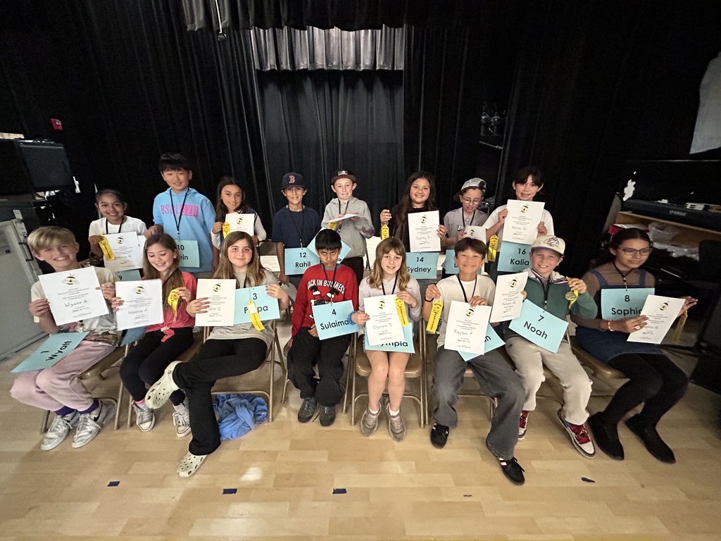 Sixteen spelling bee participants sit on stage holding certificates and ribbons, smiling proudly at the conclusion of the event.