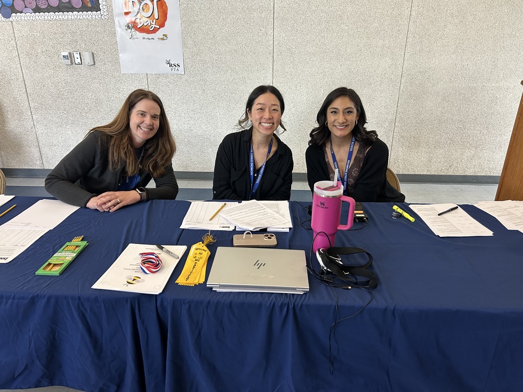 Three staff members sit behind a blue table with medals, ribbons, and paperwork, smiling at the camera during the spelling bee event.