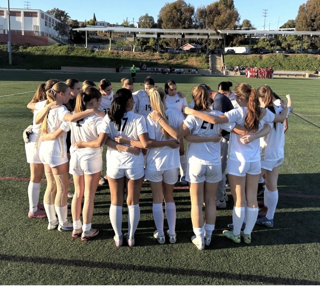 Female soccer players in white uniforms huddling together on a sunlit field before a game.